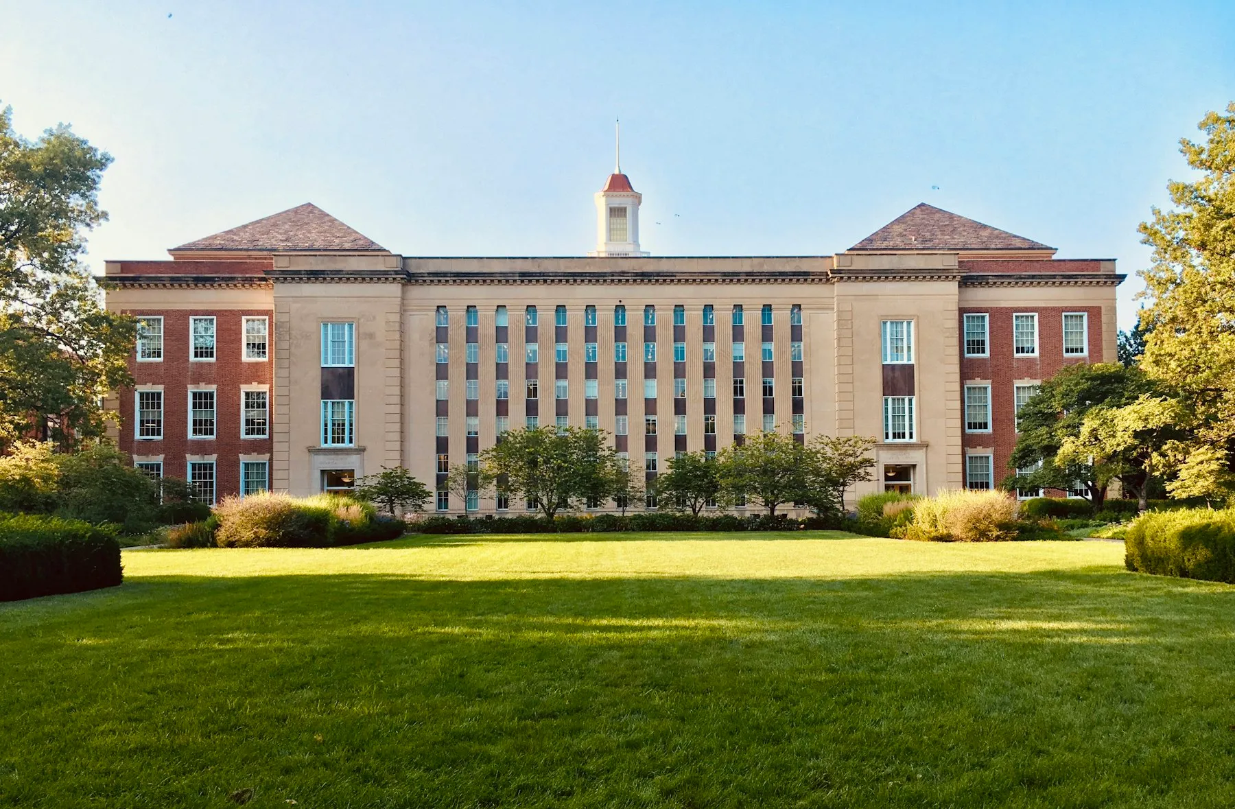 Red-brick and stone college building behind a sunlit lawn